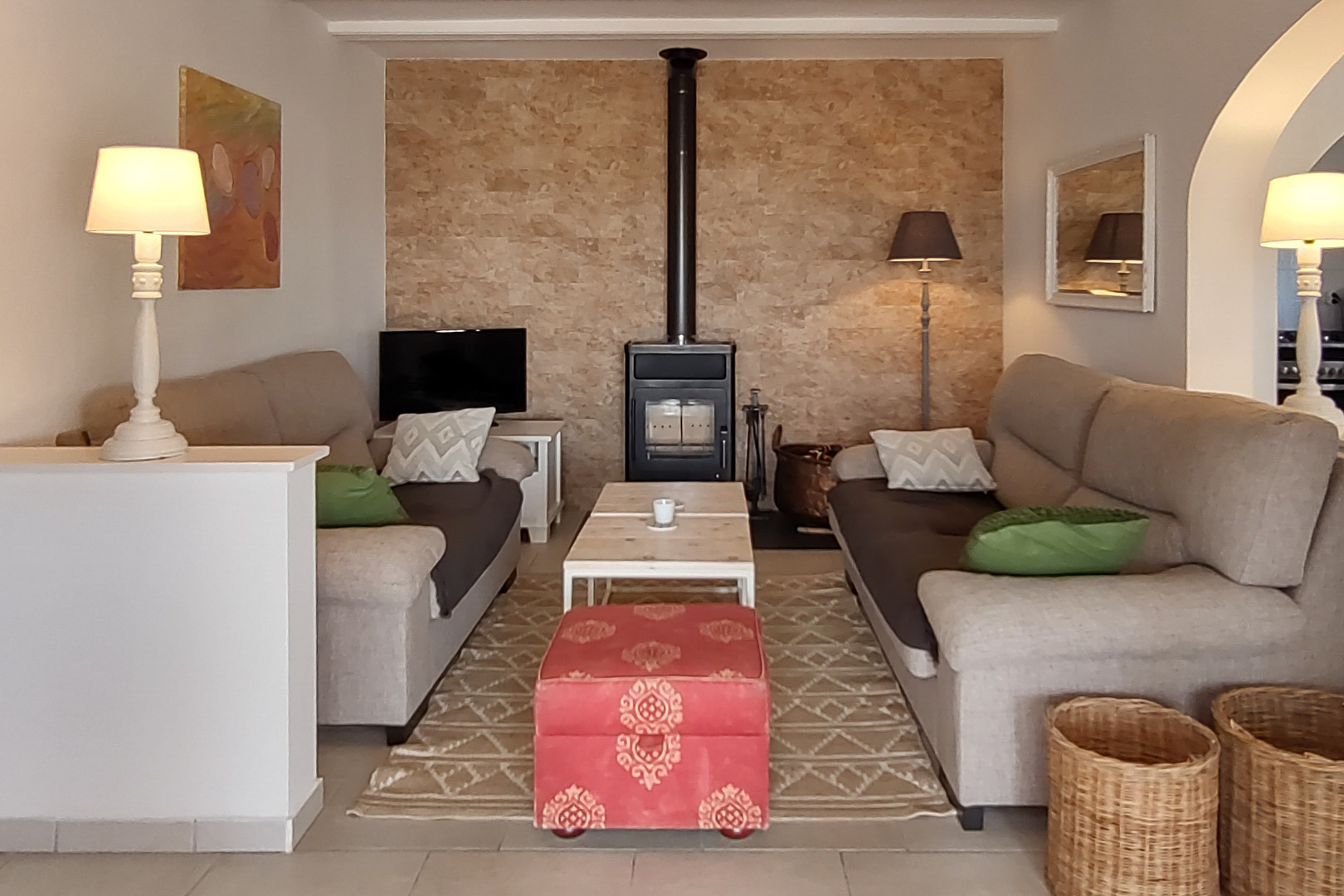 Salon chaleureux avec poêle à bois sur mur en pierre naturelle, canapés face à face, table basse en bois et pouf rouge décoratif.