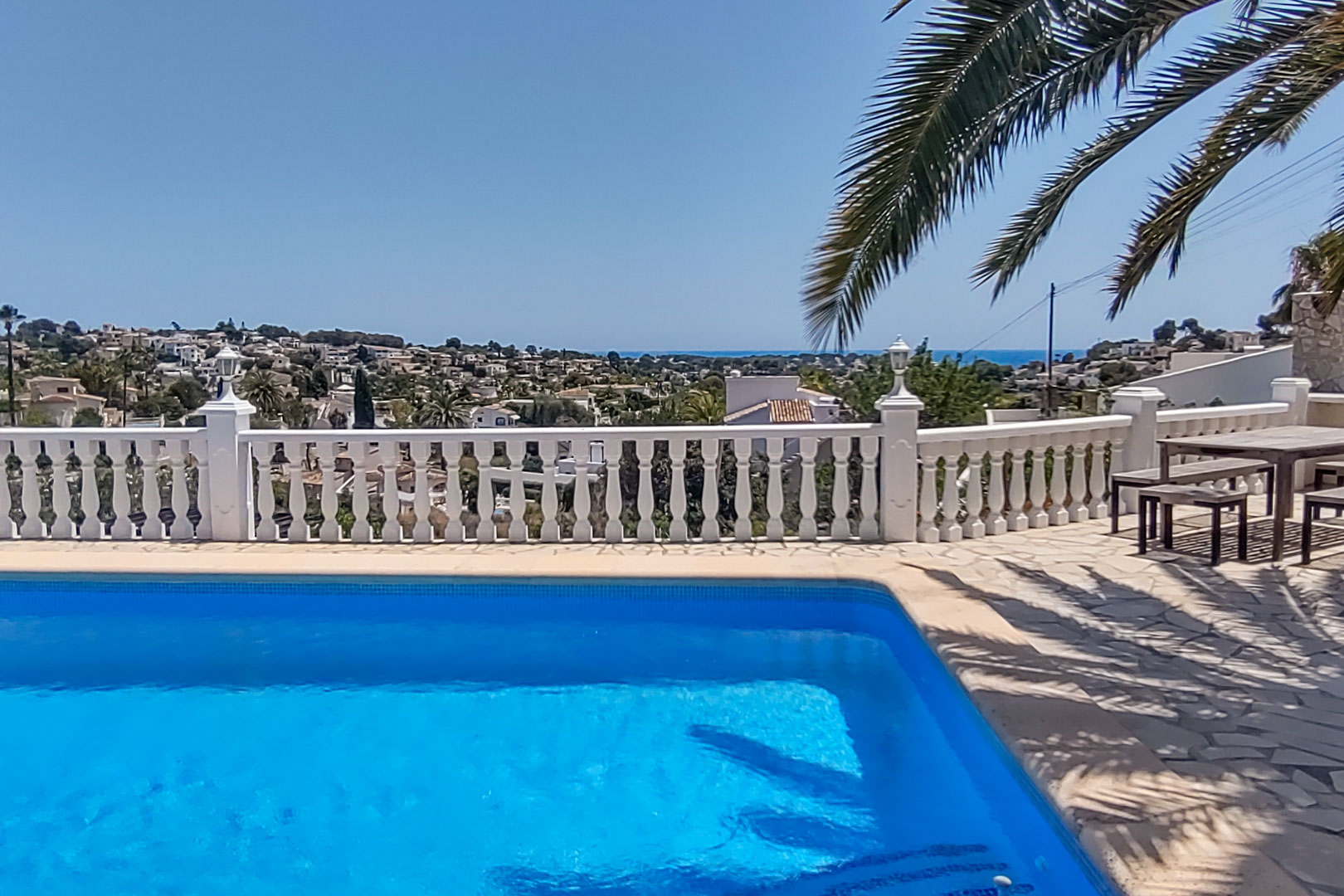 Terrasse avec piscine et balustrade blanche offrant une vue panoramique sur la mer et la vallée, avec un coin repas extérieur en bois.