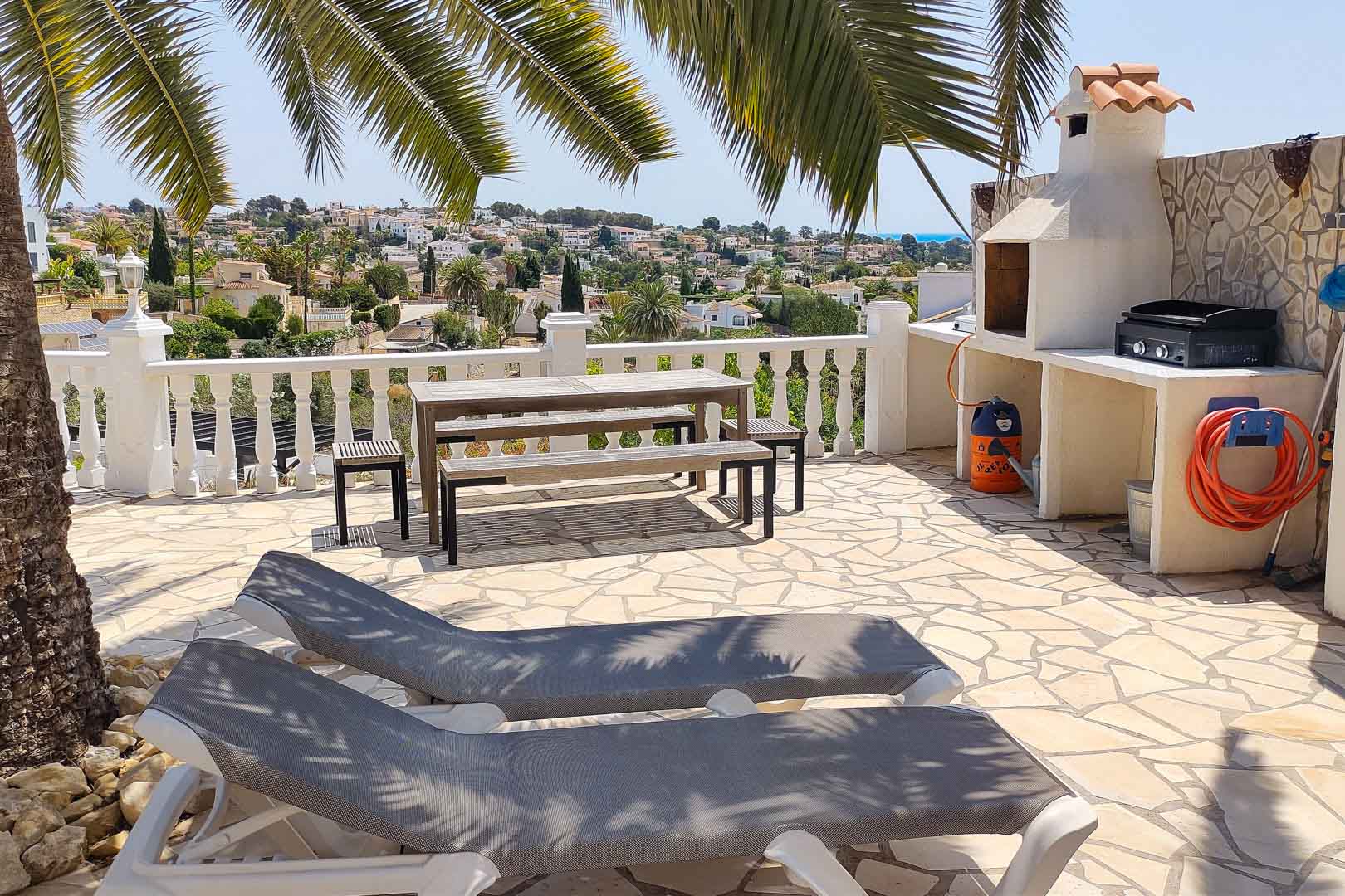 Terrasse extérieure en pierre avec barbecue maçonné, table de pique-nique en bois et chaises longues, offrant une vue dégagée sur la vallée et la mer.