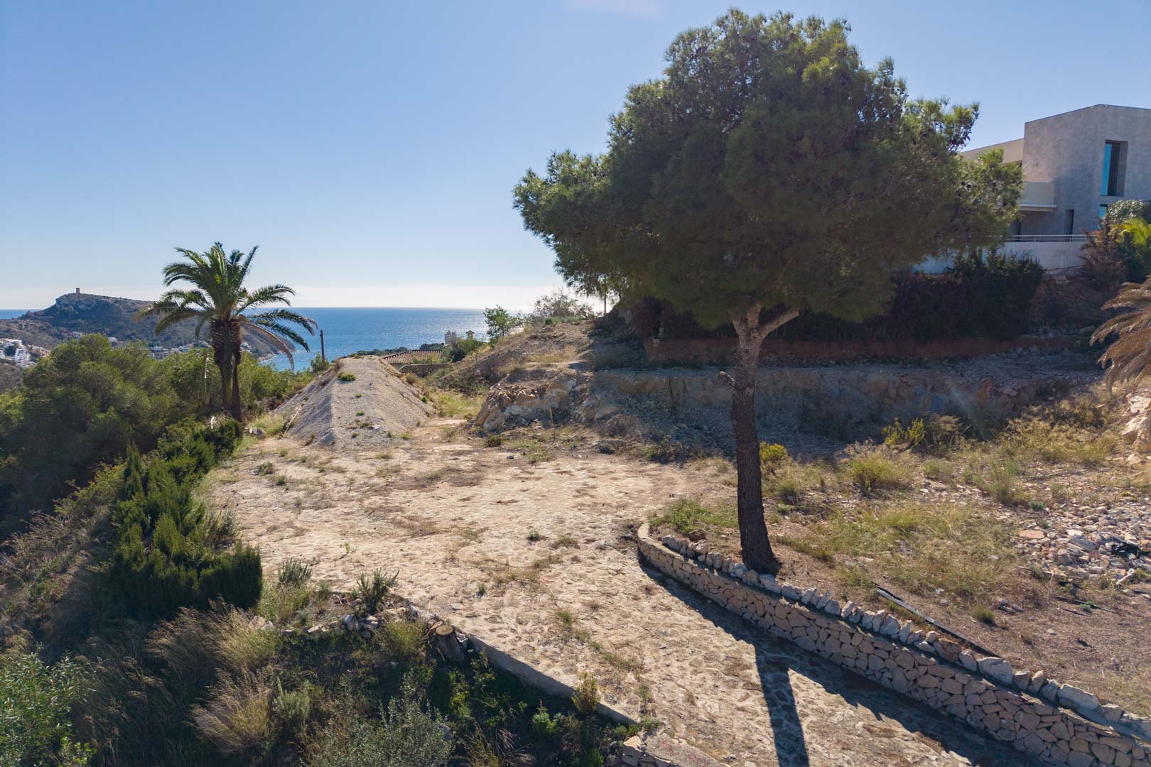 Vue extérieure du terrain avec chemin en pierre naturelle, pins, palmiers et vue panoramique sur la mer Méditerranée à Moraira.