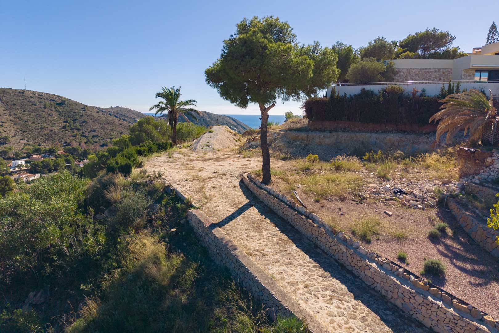 Vue panoramique depuis le terrain avec un chemin pavé et des murs en pierre sèche, entourée de végétation méditerranéenne et vue sur mer.