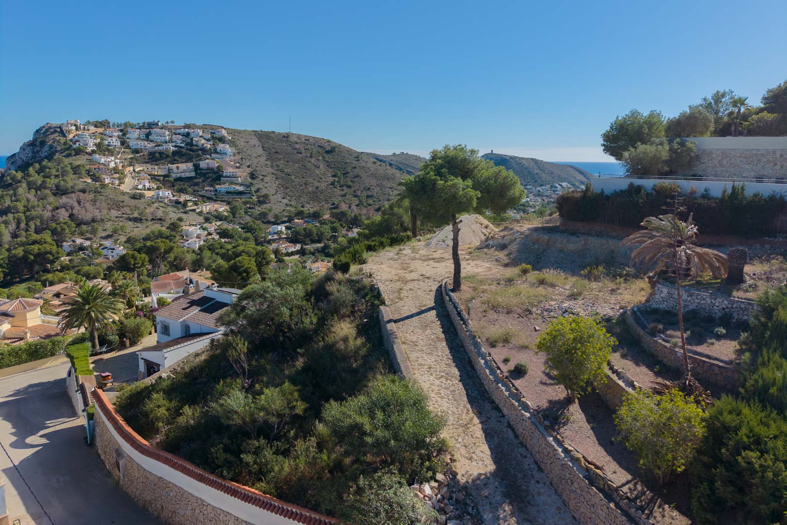 Vue surélevée du terrain avec des murs en pierre naturelle et une végétation méditerranéenne, offrant une vue panoramique sur la vallée et la mer à Moraira.