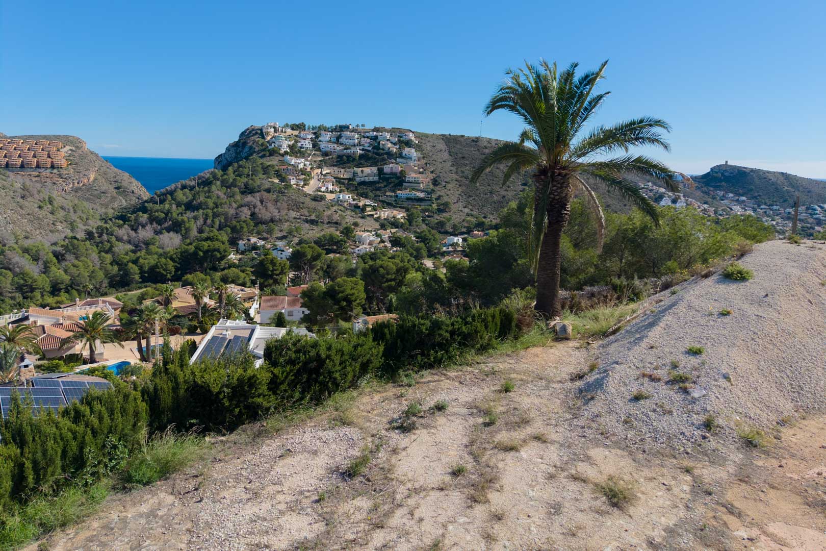 Vues panoramiques depuis le terrain surélevé sur la vallée de Moraira, avec la mer Méditerranée à l'horizon et des collines couvertes de pins et de palmiers.