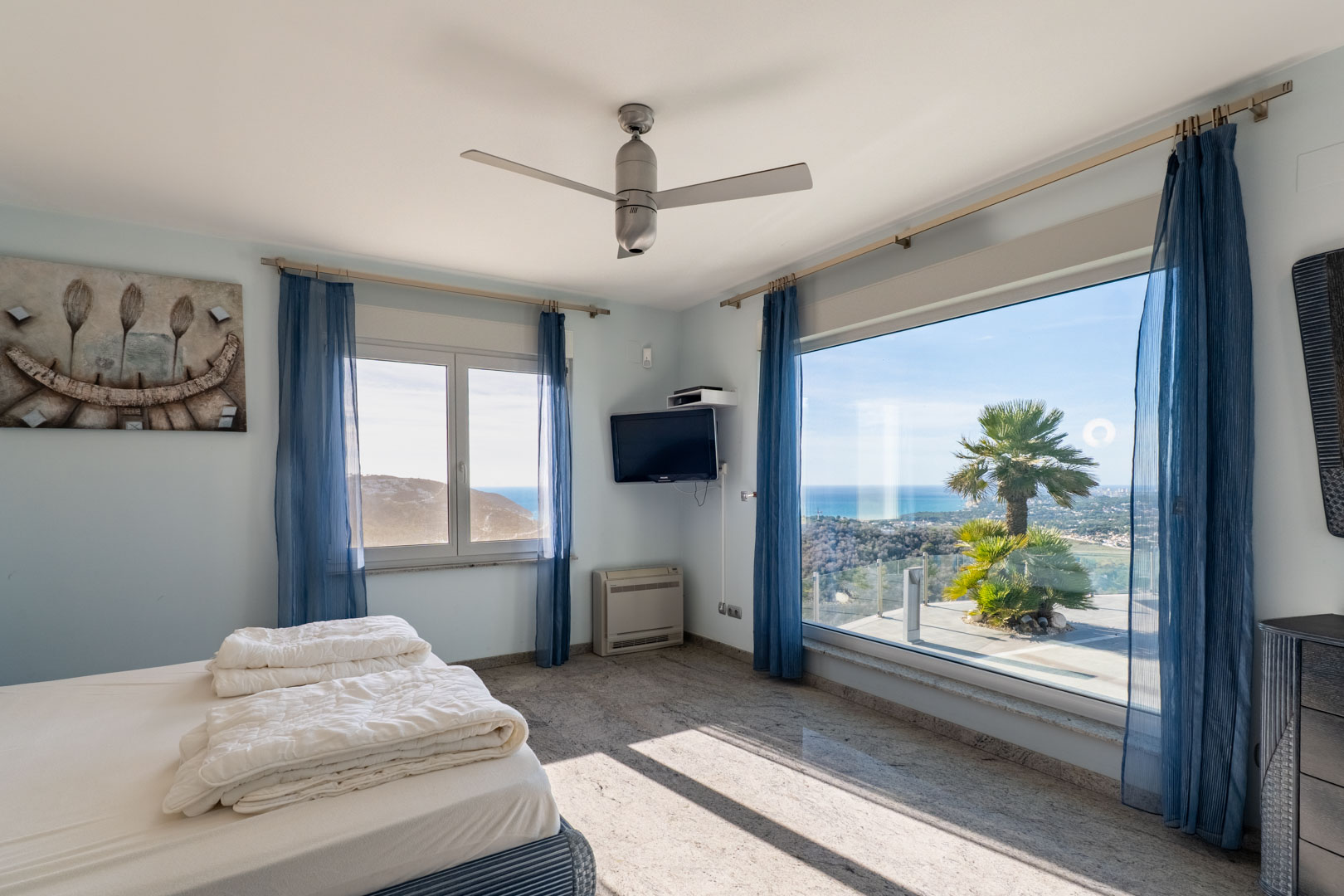 Chambre lumineuse avec sol en granit, rideaux bleus et vue sur la mer à travers de grandes fenêtres.