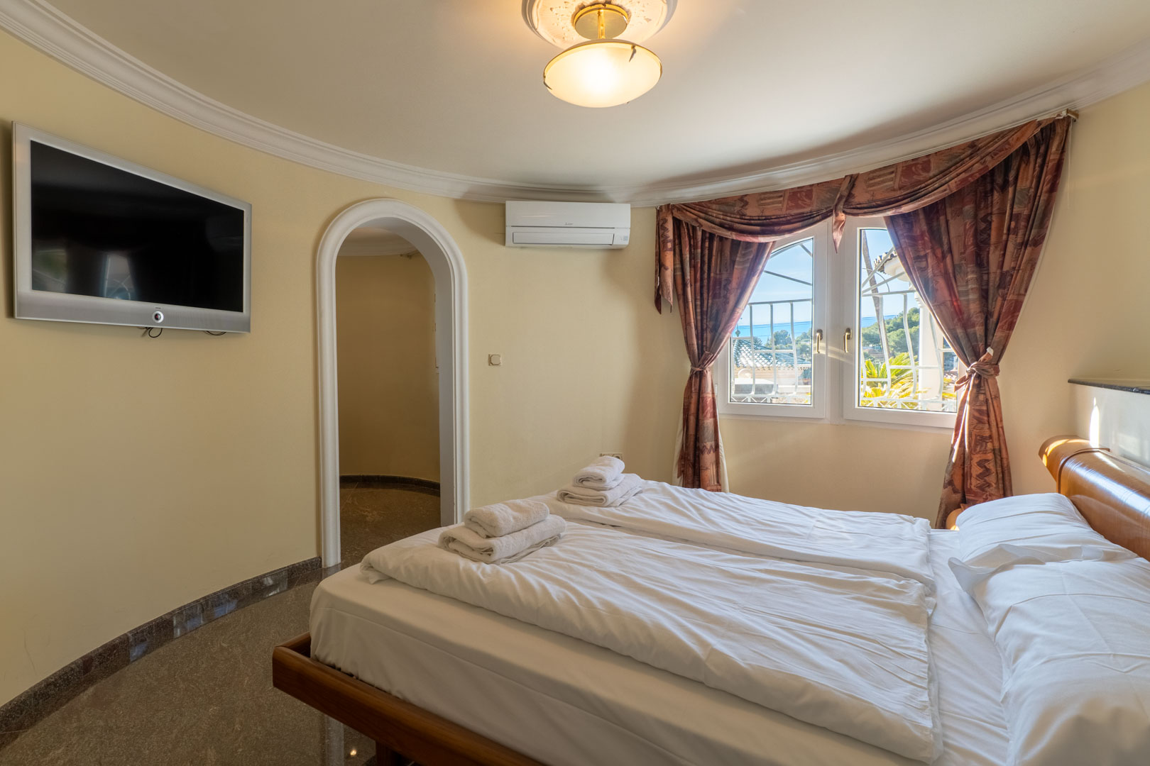 Chambre avec mur courbe, porte en arche et sol en pierre polie. Elle dispose d'une télévision murale, climatisation et fenêtre offrant une vue sur la mer.