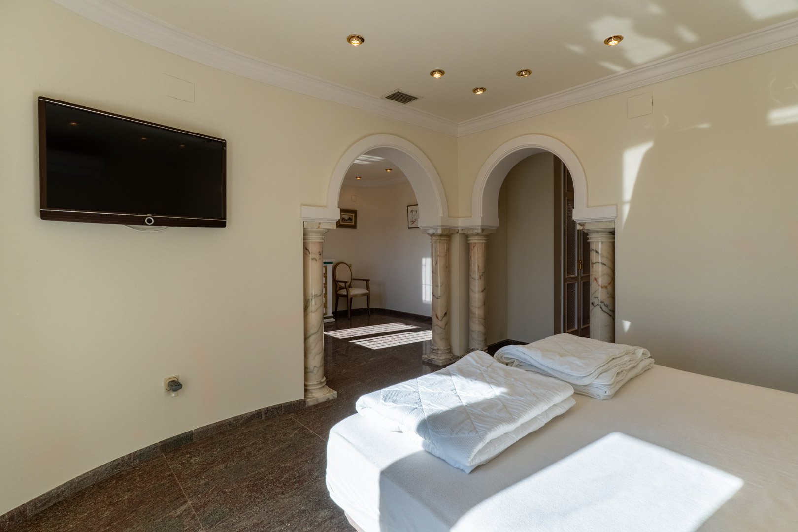 Chambre principale avec arches classiques sur colonnes en marbre, sol en pierre polie et accès à un espace privé adjacent baigné de lumière naturelle.