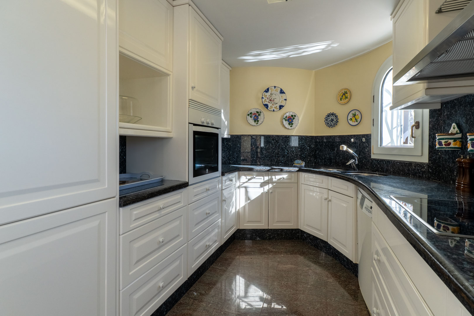 Cuisine indépendante avec meubles blancs, plan de travail en granit sombre et assiettes décoratives. Elle dispose d'une fenêtre cintrée et d'appareils intégrés.