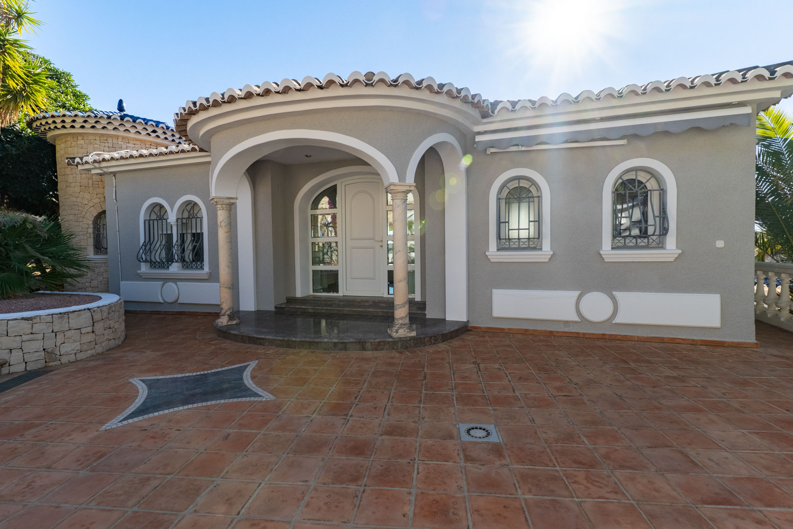 Entrée principale avec porche voûté soutenu par des colonnes en marbre, terrasse en terre cuite avec mosaïque et façade grise aux fenêtres cintrées ferronnées.