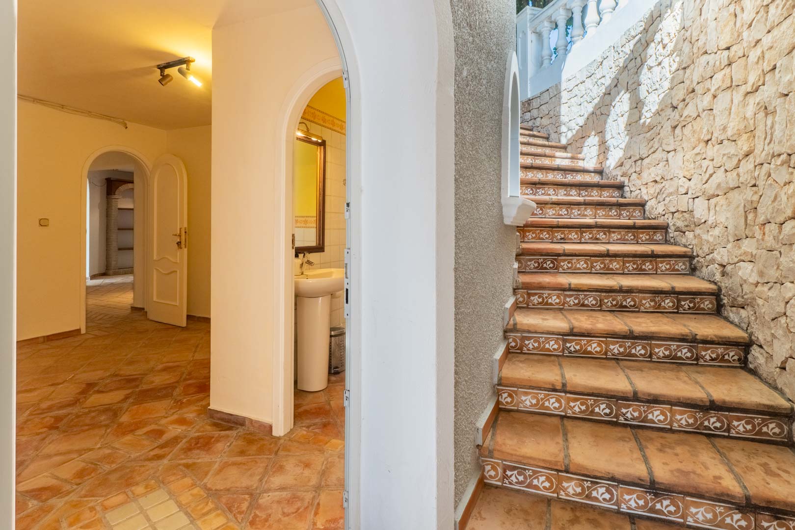 Escalier extérieur avec contremarches décorées et mur en pierre, menant à un couloir intérieur avec sol en terre cuite et passages voûtés.