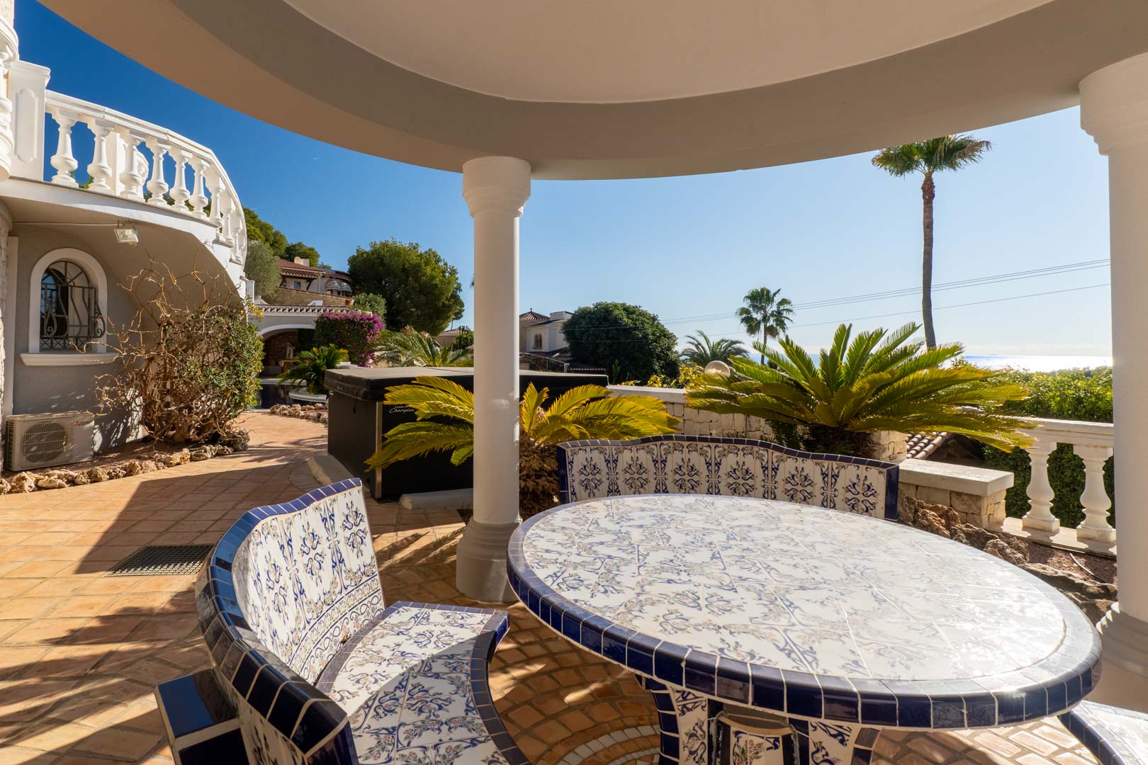 Espace dînatoire extérieur sous porche, doté d'une table et de bancs en céramique traditionnelle bleue et blanche, offrant une vue sur le jardin et la mer.