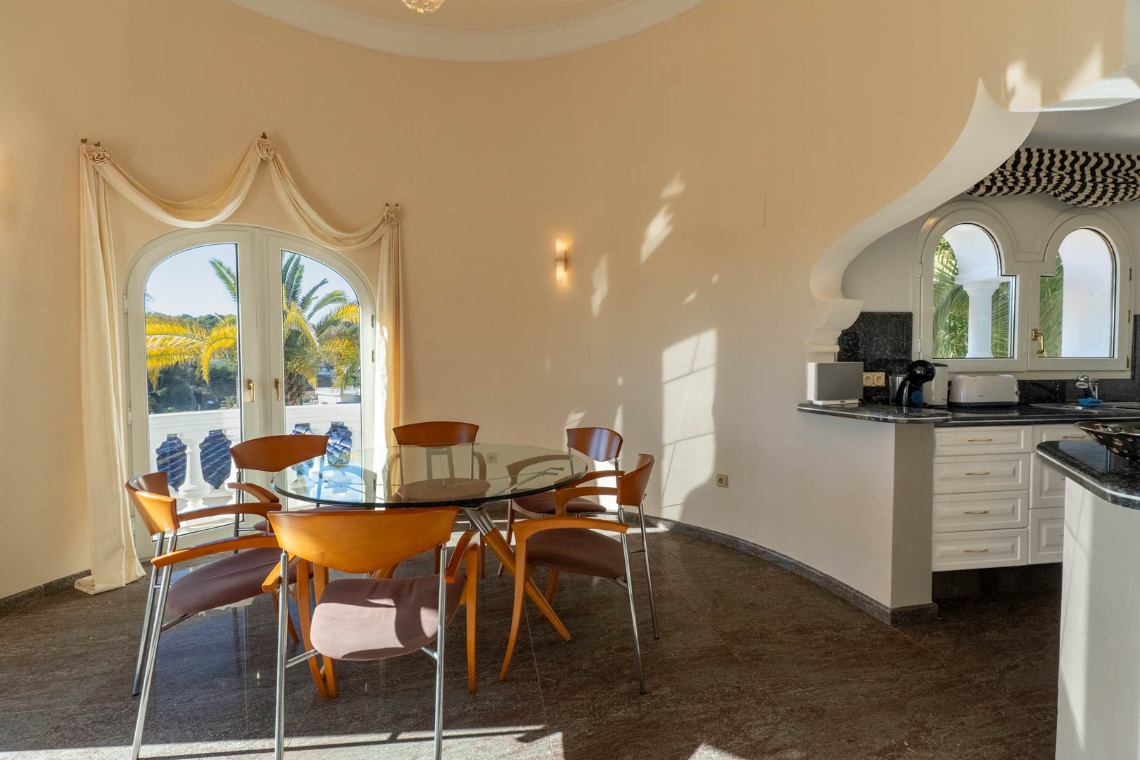Espace repas circulaire avec table en verre et chaises en bois, attenant à une cuisine lumineuse avec plans de travail sombres et accès au balcon balustré.