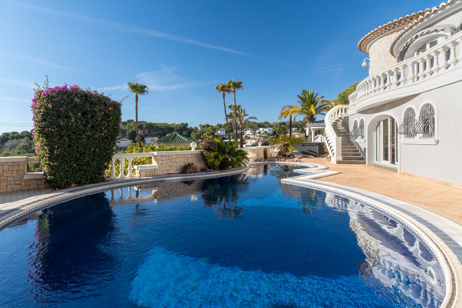 Piscine aux formes organiques avec mosaïque bleue, entourée d'une terrasse en terre cuite et de murs en pierre, à côté de la façade blanche aux fenêtres cintrées et balustrades.