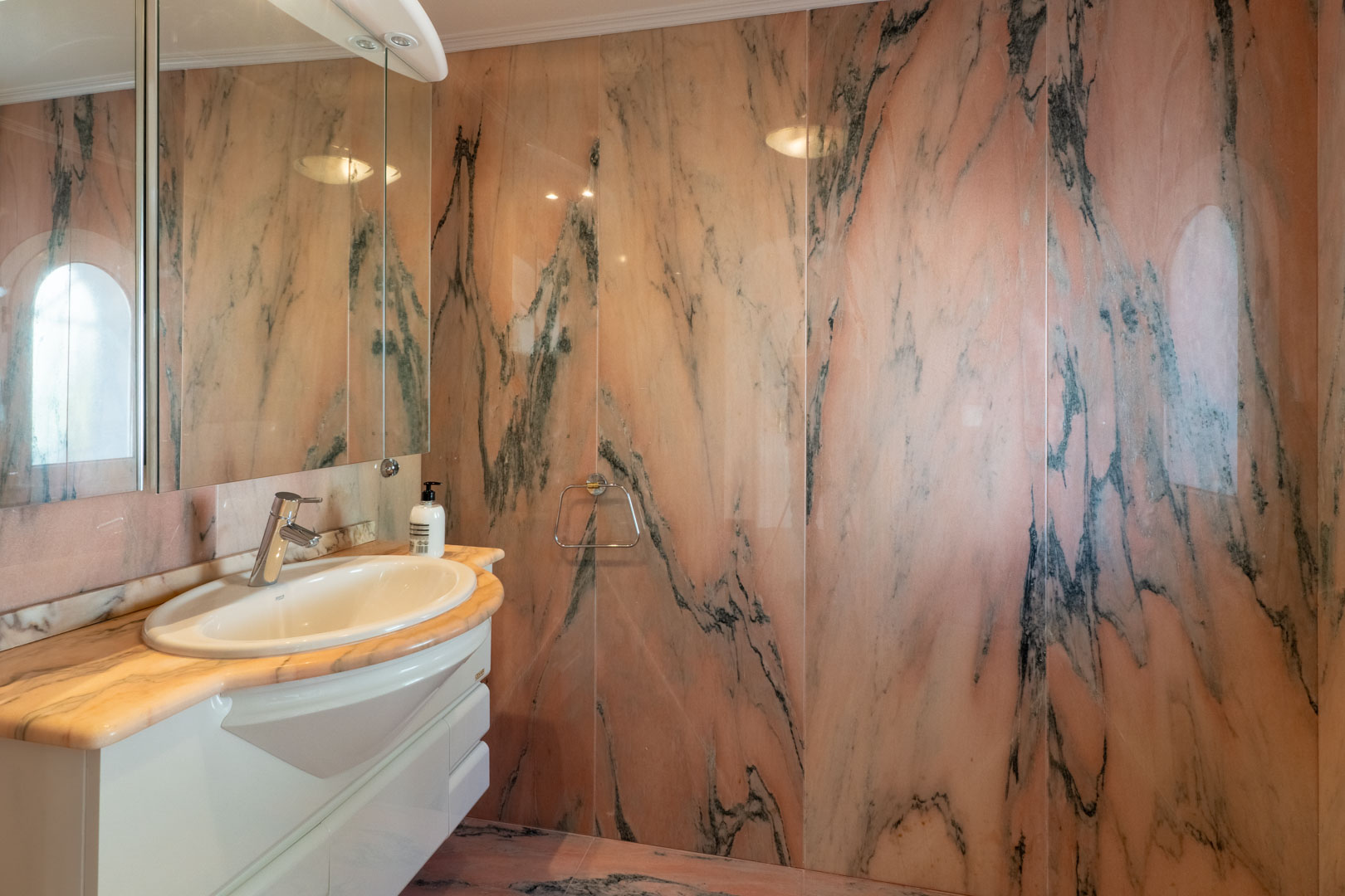 Salle de bain avec revêtement mural complet en marbre rose veiné. Elle dispose d'un plan de travail en pierre assorti, d'un meuble vasque blanc et d'un grand miroir.