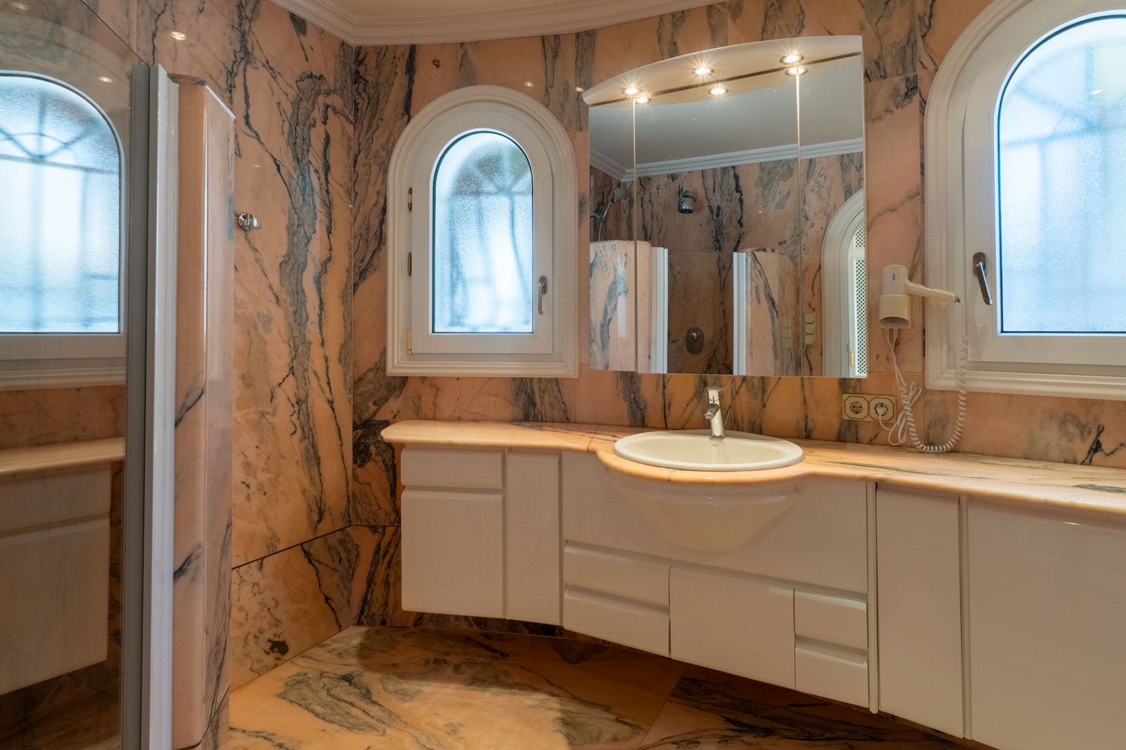 Salle de bain revêtue de marbre rose veiné, comprenant un meuble vasque blanc, un miroir avec spots et des fenêtres cintrées en verre dépoli.