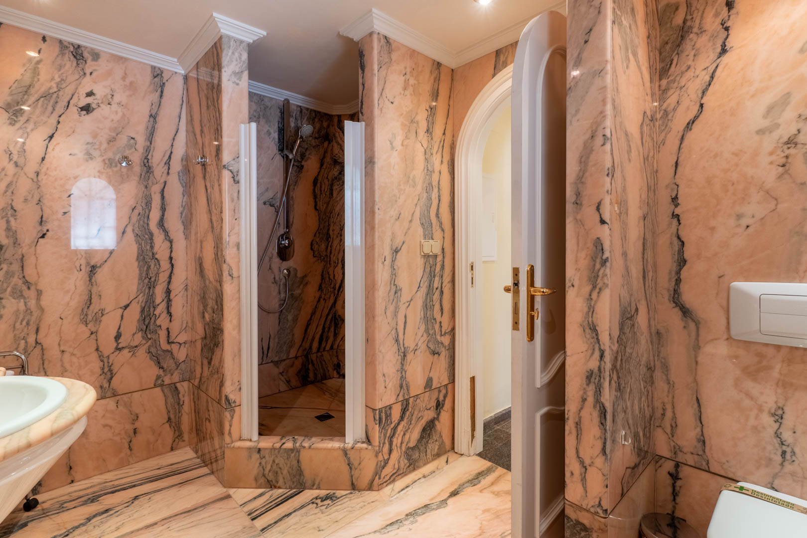 Salle de bains revêtue de marbre rose veiné, comprenant une douche à l'italienne, des moulures classiques et une porte cintrée avec quincaillerie dorée.