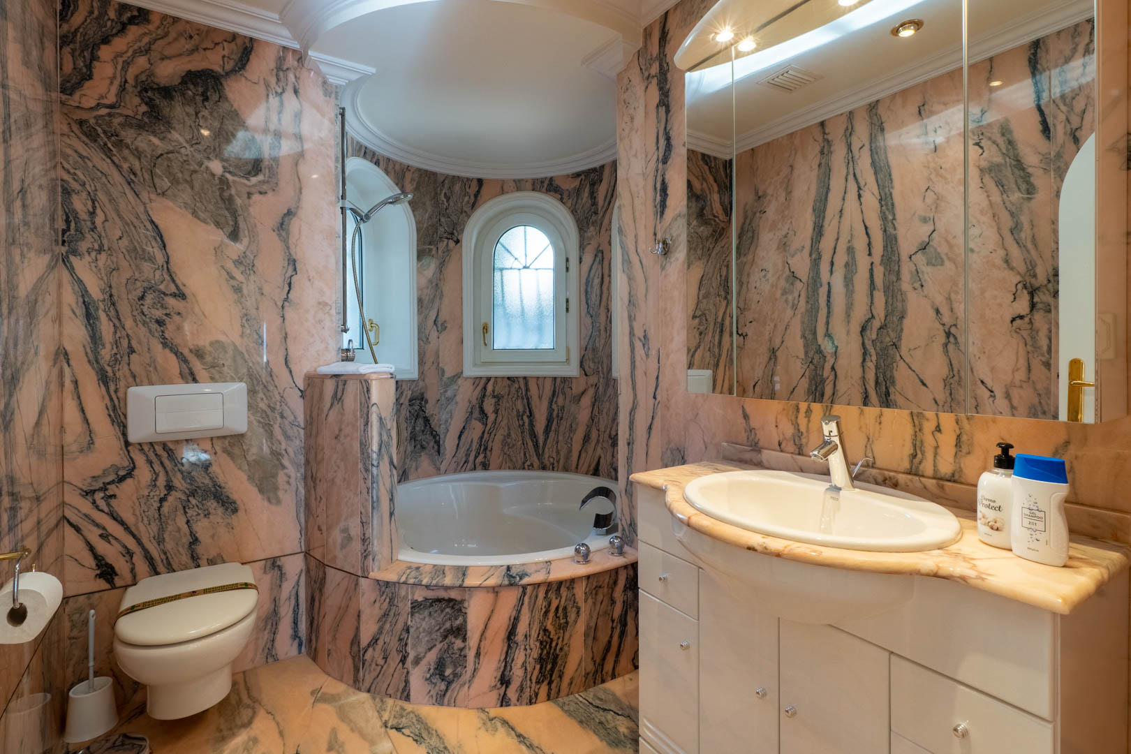 Salle de bains revêtue de marbre veiné rose et gris, comprenant une baignoire encastrée dans une alcôve courbe, des fenêtres en plein cintre et un meuble vasque blanc.