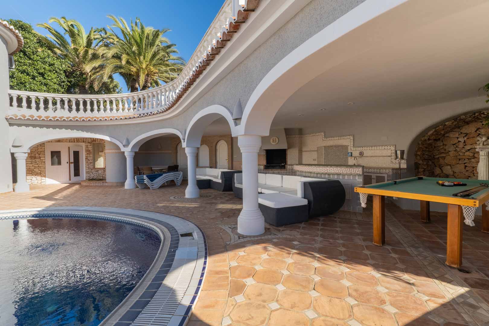 Terrasse couverte avec arches au bord de la piscine, comprenant un salon extérieur, un billard et une cuisine d'été avec finitions en céramique.