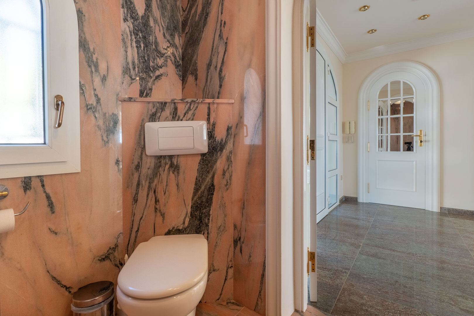 Toilettes invités avec murs en marbre rose veiné, WC suspendu et vue sur un couloir au sol en pierre polie avec des portes blanches cintrées.
