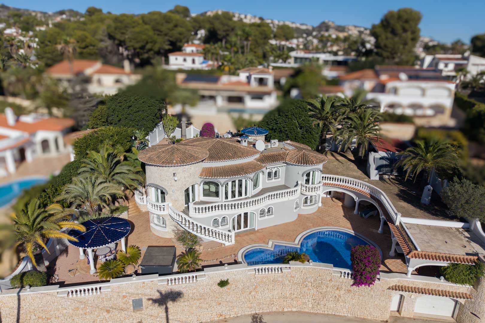 Vue aérienne d'une villa méditerranéenne avec tour en pierre, piscine incurvée, terrasses à balustrades blanches et jardin paysager agrémenté de palmiers à Benissa.