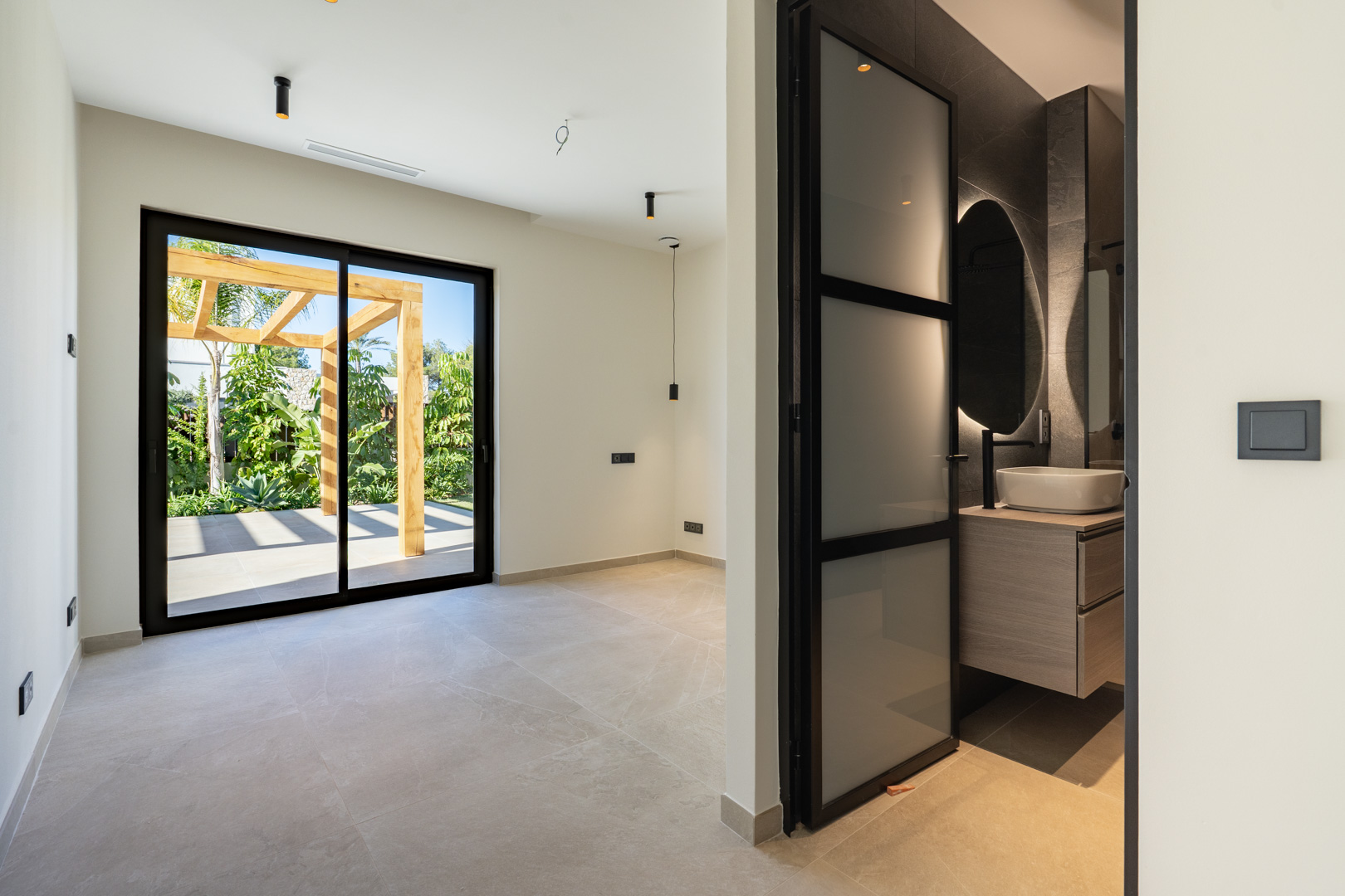 Chambre avec accès terrasse et pergola en bois. Comprend une salle de bain attenante avec vasque à poser, miroir rétroéclairé et menuiserie noire élégante.