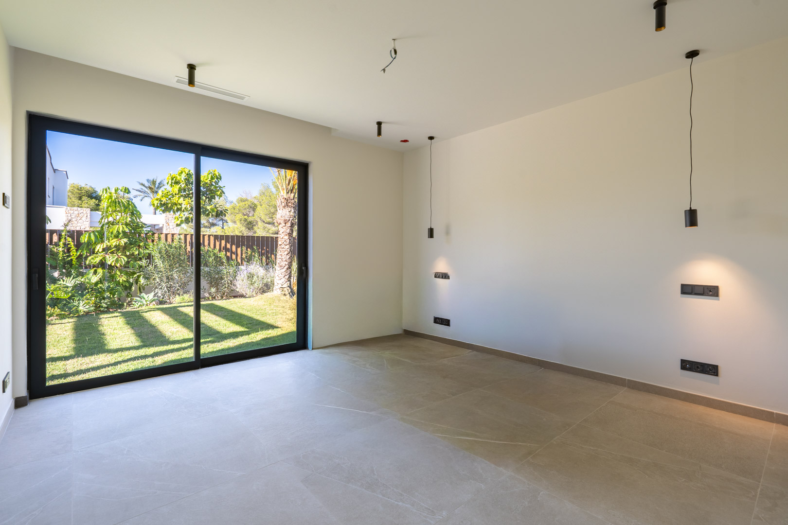 Chambre avec sol en grès cérame et grande baie vitrée coulissante ouvrant sur le jardin. Éclairage minimaliste noir et système de climatisation intégré au plafond.