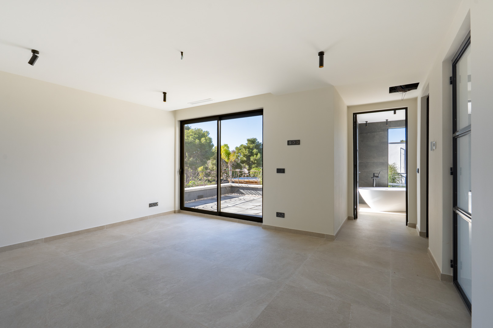 Chambre principale avec accès à une terrasse privée, carrelage grand format et salle de bain attenante avec baignoire îlot et murs gris.