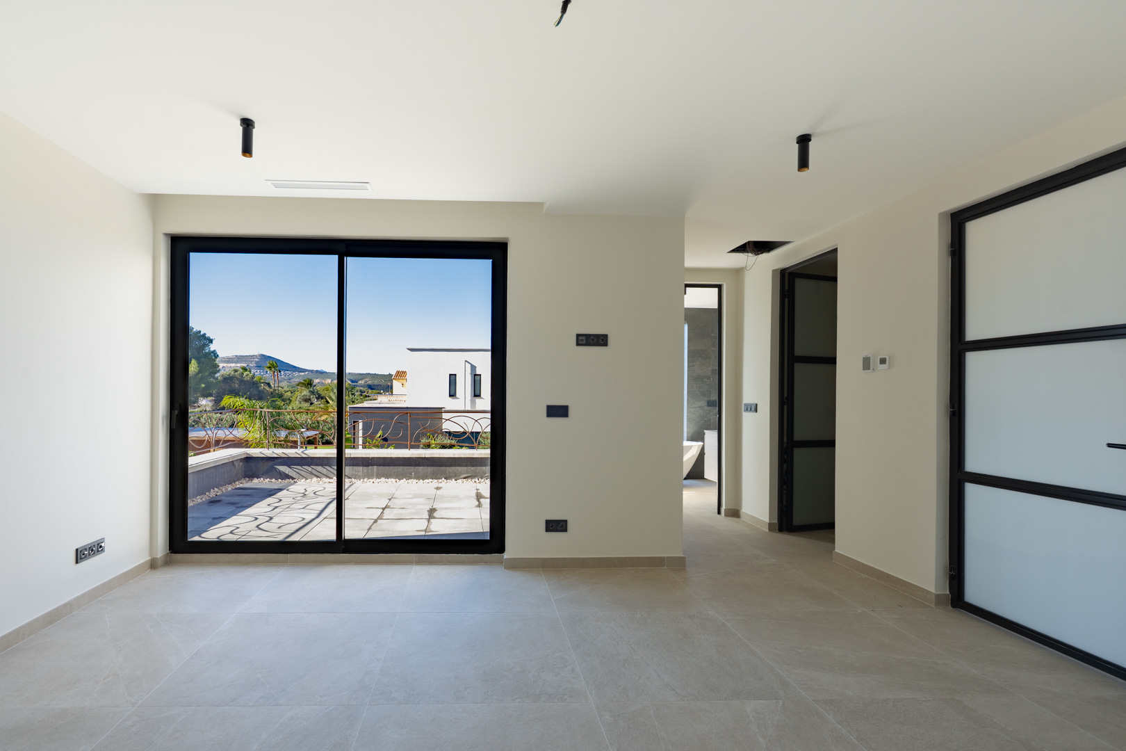 Chambre principale lumineuse avec accès terrasse, sol en grès cérame et menuiseries noires. Aperçu de la salle de bain attenante avec baignoire îlot.