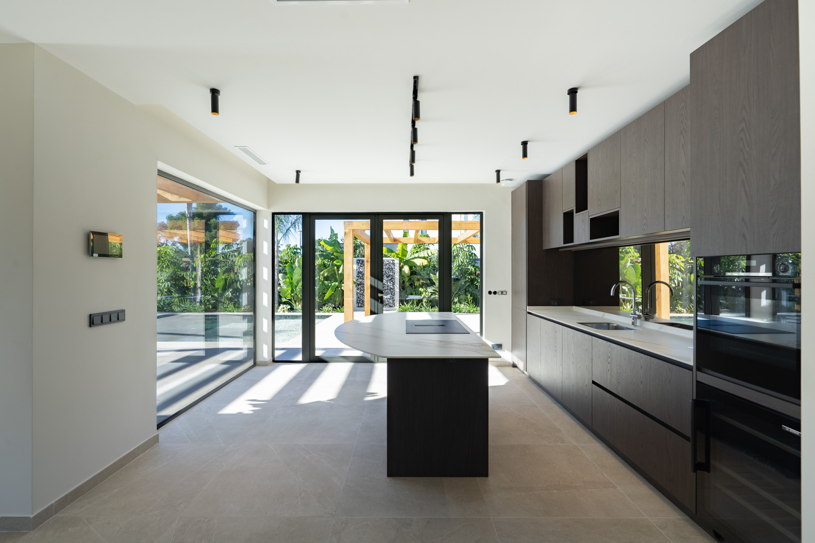 Cuisine moderne avec îlot central en pierre, meubles en bois sombre et électroménager intégré. Les baies vitrées s'ouvrent sur le jardin méditerranéen et la piscine.