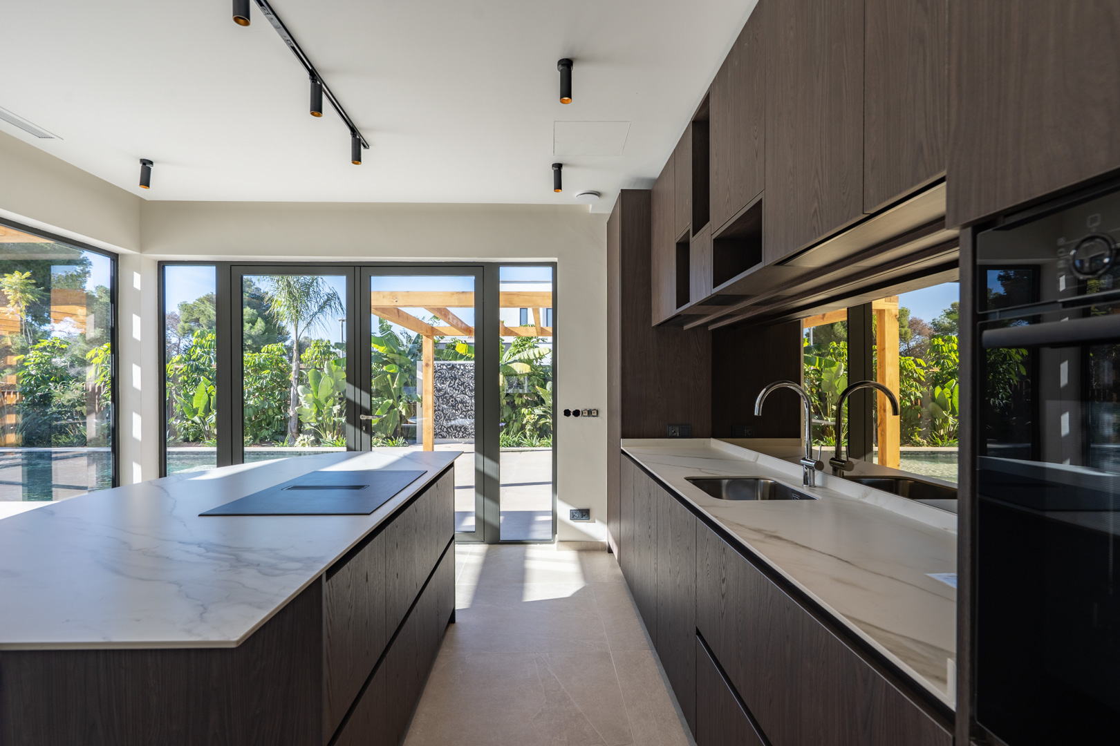 Cuisine moderne avec îlot central, plans de travail effet marbre et meubles en bois sombre. Les baies vitrées s'ouvrent sur le jardin méditerranéen et la piscine.