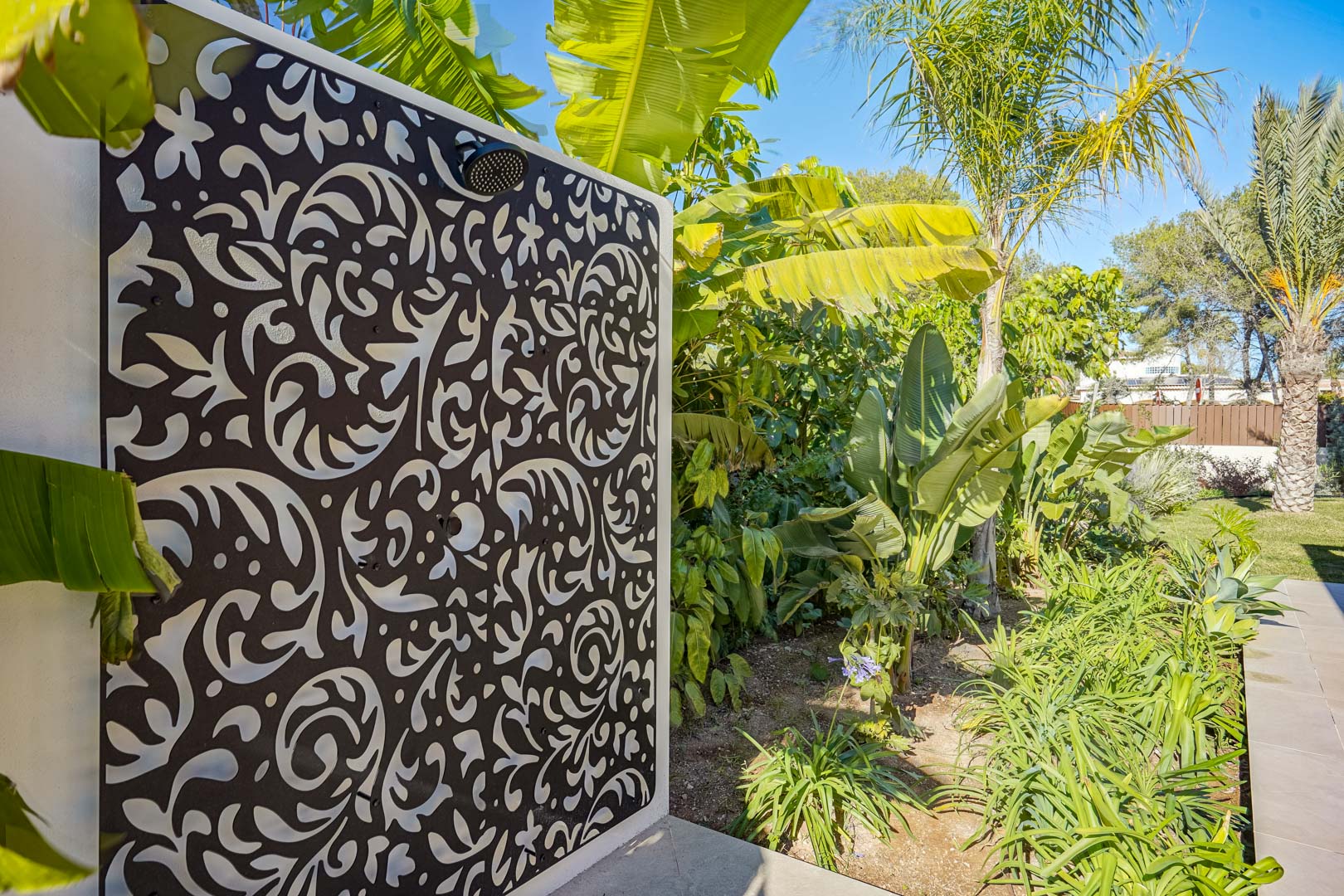 Douche extérieure avec panneau mural décoratif découpé au laser, nichée dans un jardin méditerranéen luxuriant aux accents tropicaux.