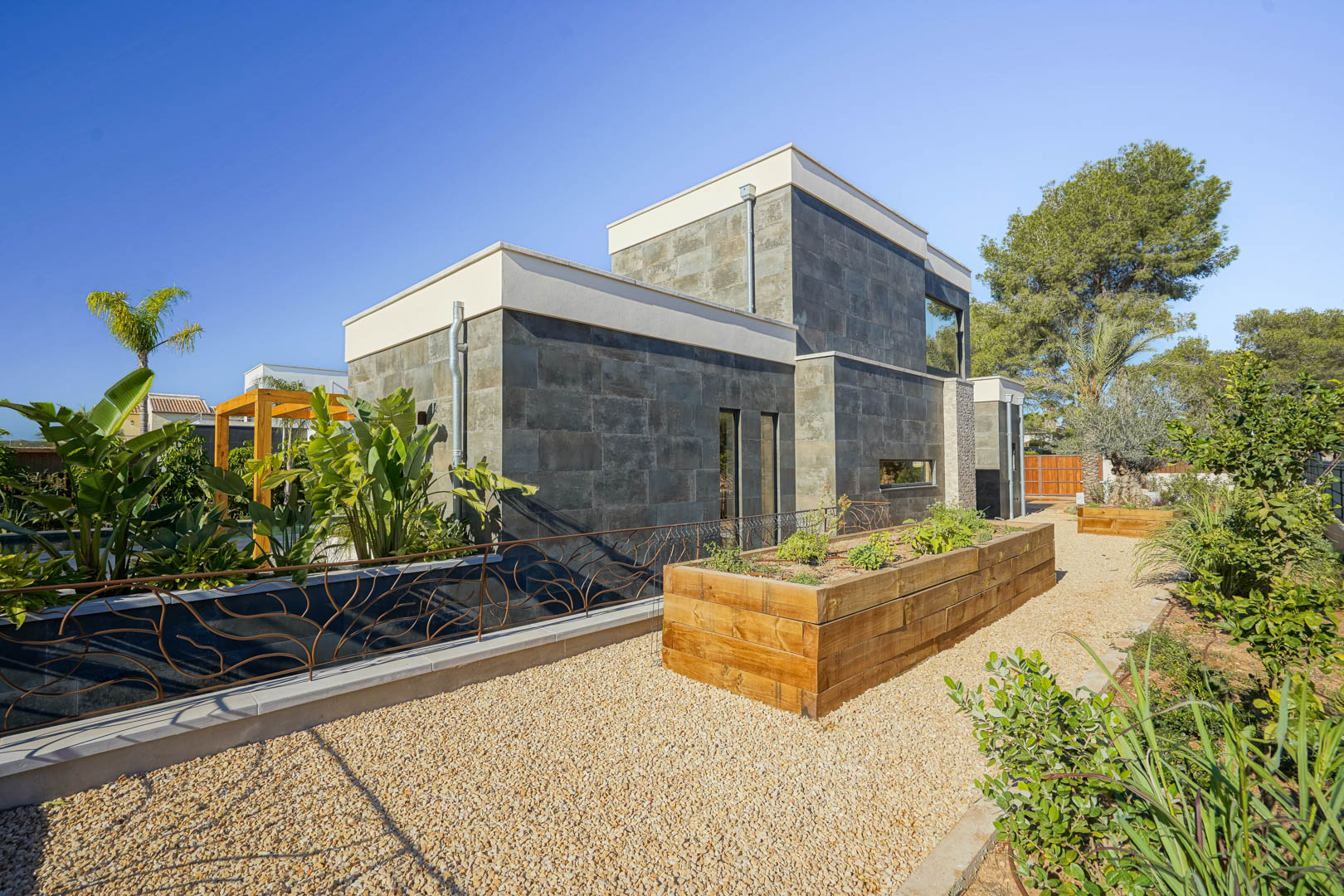 Façade contemporaine en pierre grise avec des jardinières en bois surélevées et des allées de gravier dans un jardin méditerranéen.