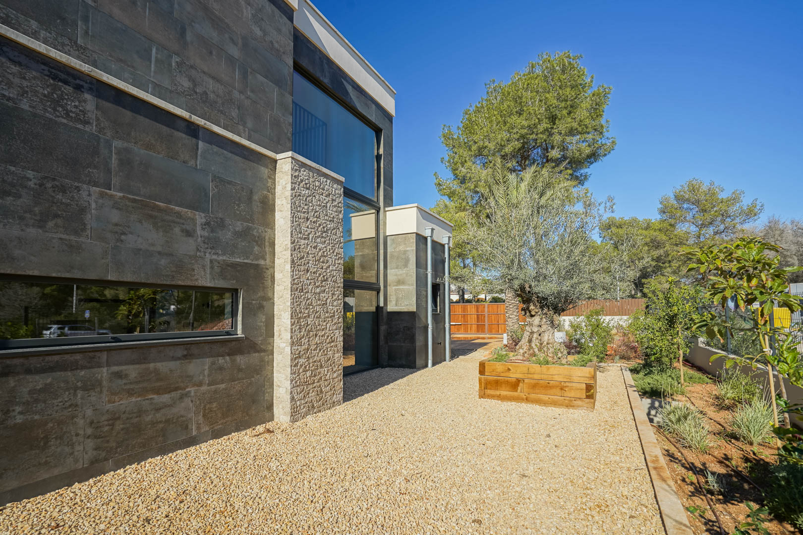Façade moderne avec revêtement en pierre sombre, accents en pierre naturelle texturée, grandes baies vitrées et jardin méditerranéen avec un olivier ancien.