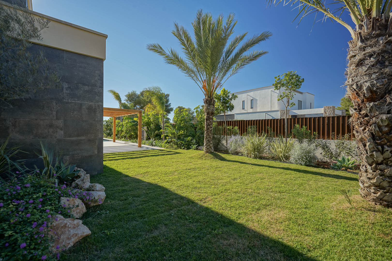 Jardin méditerranéen avec pelouse naturelle, palmiers et plantes locales, bordé par une façade en pierre sombre et une pergola en bois d'iroko.