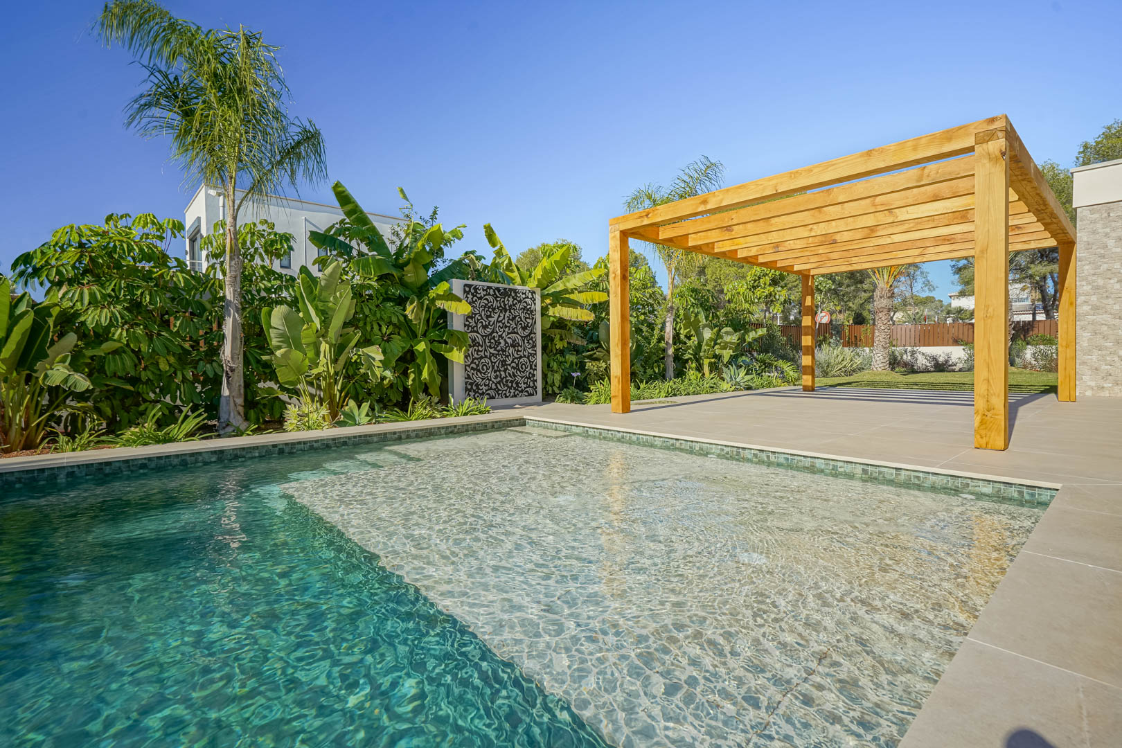Piscine de style Bali avec zone de repos, entourée d'une terrasse en grès cérame et d'une pergola en bois d'iroko au cœur d'un jardin méditerranéen.