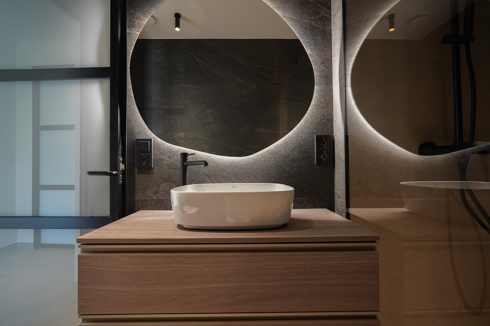 Salle de bain contemporaine avec vasque à poser sur meuble en bois, miroir rond rétroéclairé et parois en pierre sombre avec robinetterie noire mate.