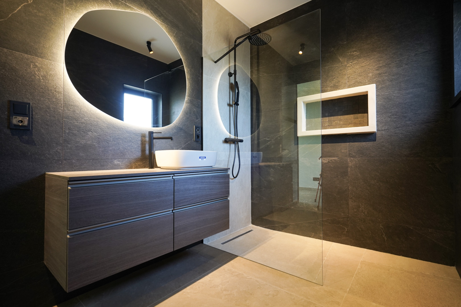 Salle de bain design avec murs en pierre sombre, vasque à poser et miroir organique rétroéclairé. Douche à l'italienne avec robinetterie noire et paroi en verre.