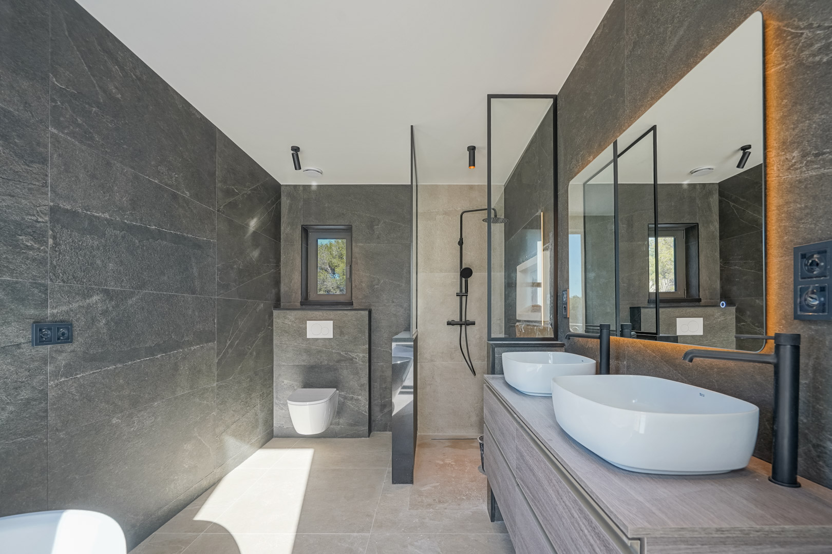 Salle de bain moderne avec carrelage effet pierre grise, double vasque sur meuble en bois, robinetterie noire mate et douche à l'italienne avec paroi vitrée.