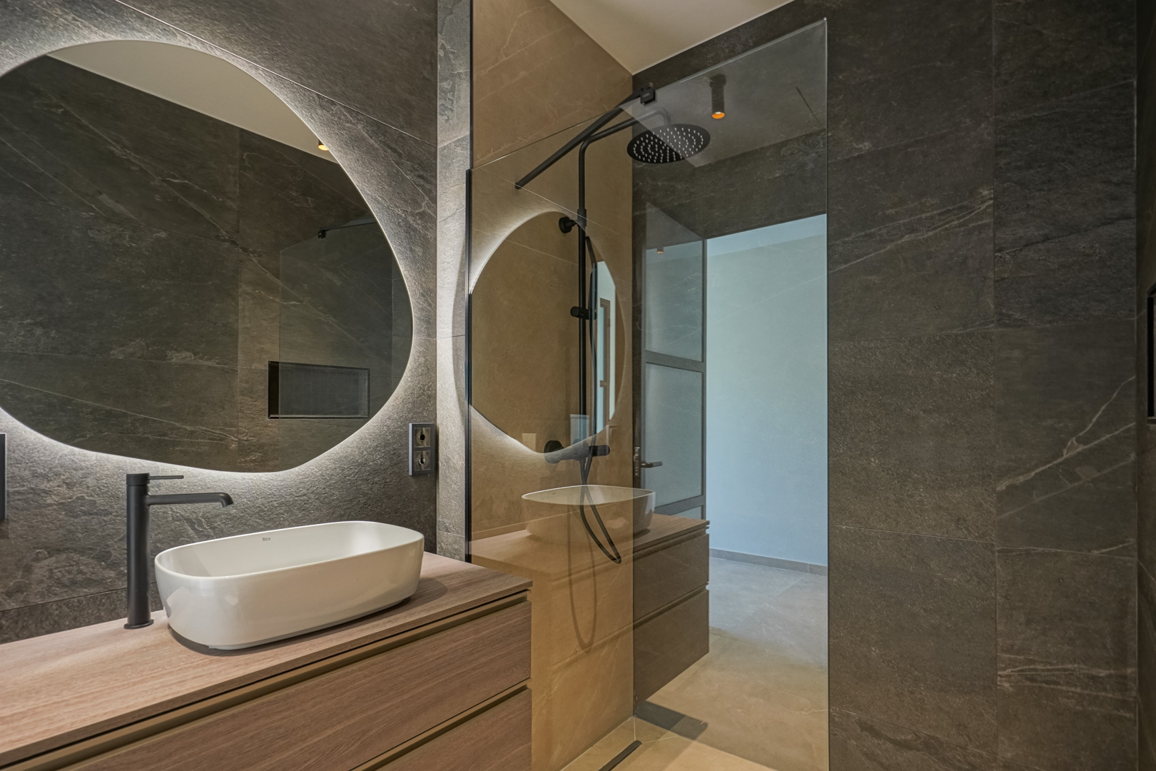 Salle de bain moderne avec carrelage effet pierre sombre, meuble vasque en bois et miroir circulaire rétroéclairé à côté d'une douche à l'italienne.