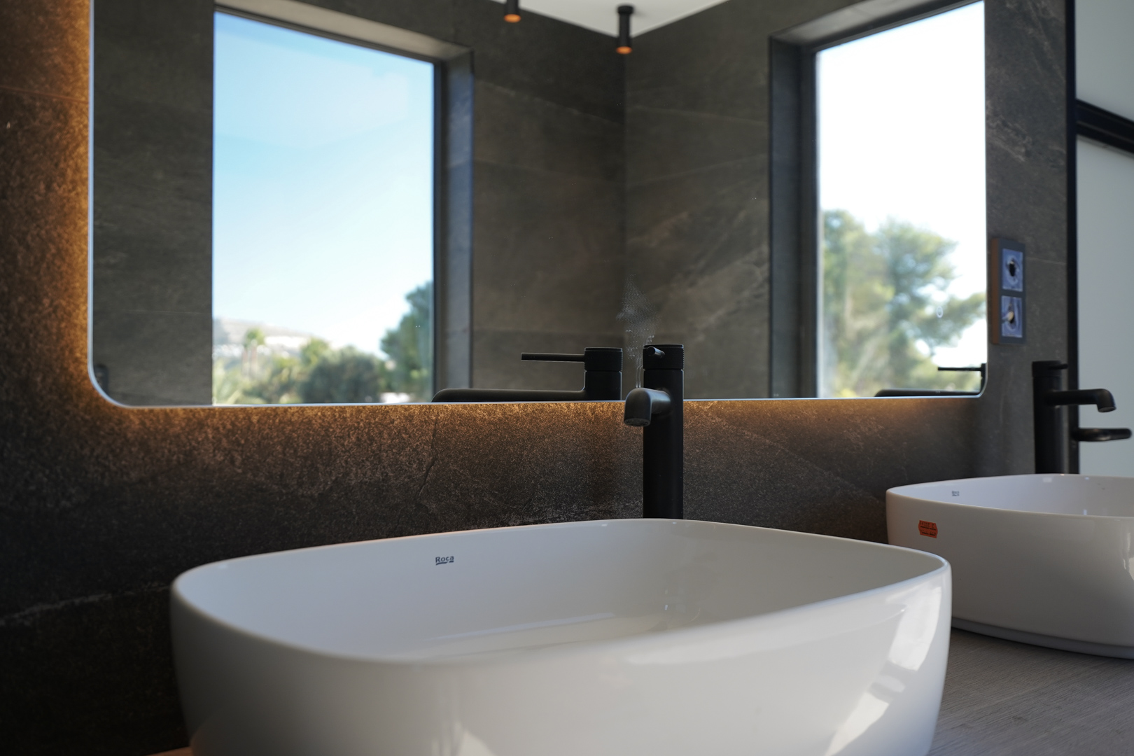 Salle de bain moderne avec vasques Roca, robinetterie noire mate et miroir rétroéclairé sur un mur en pierre sombre reflétant l'extérieur.