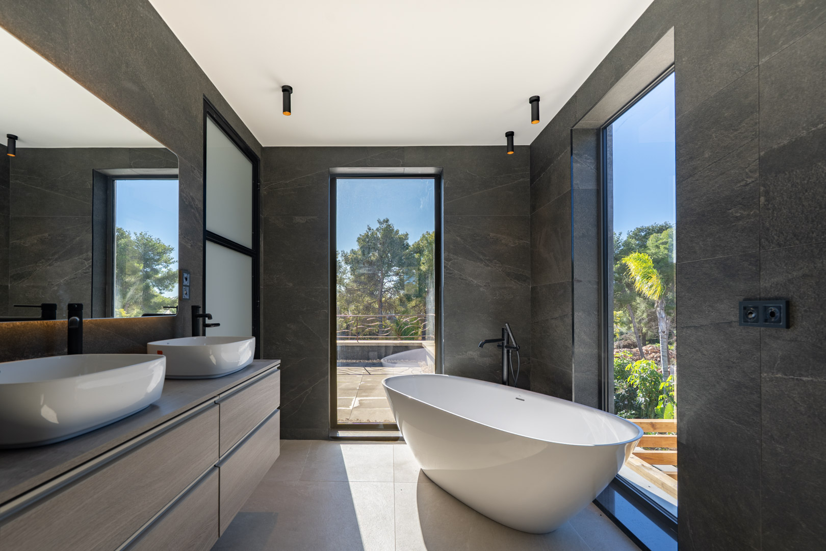 Salle de bain principale avec baignoire îlot, double vasque et grandes baies vitrées offrant une vue sur la verdure. Murs en pierre sombre et robinetterie noire élégante.