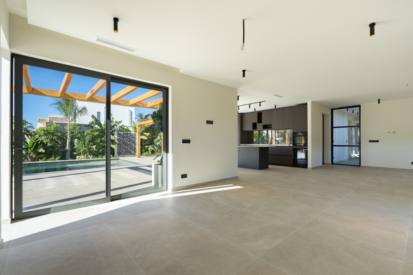 Séjour lumineux avec carrelage en grès cérame et grandes baies vitrées ouvrant sur la piscine et la pergola. Cuisine moderne intégrée en bois sombre au second plan.
