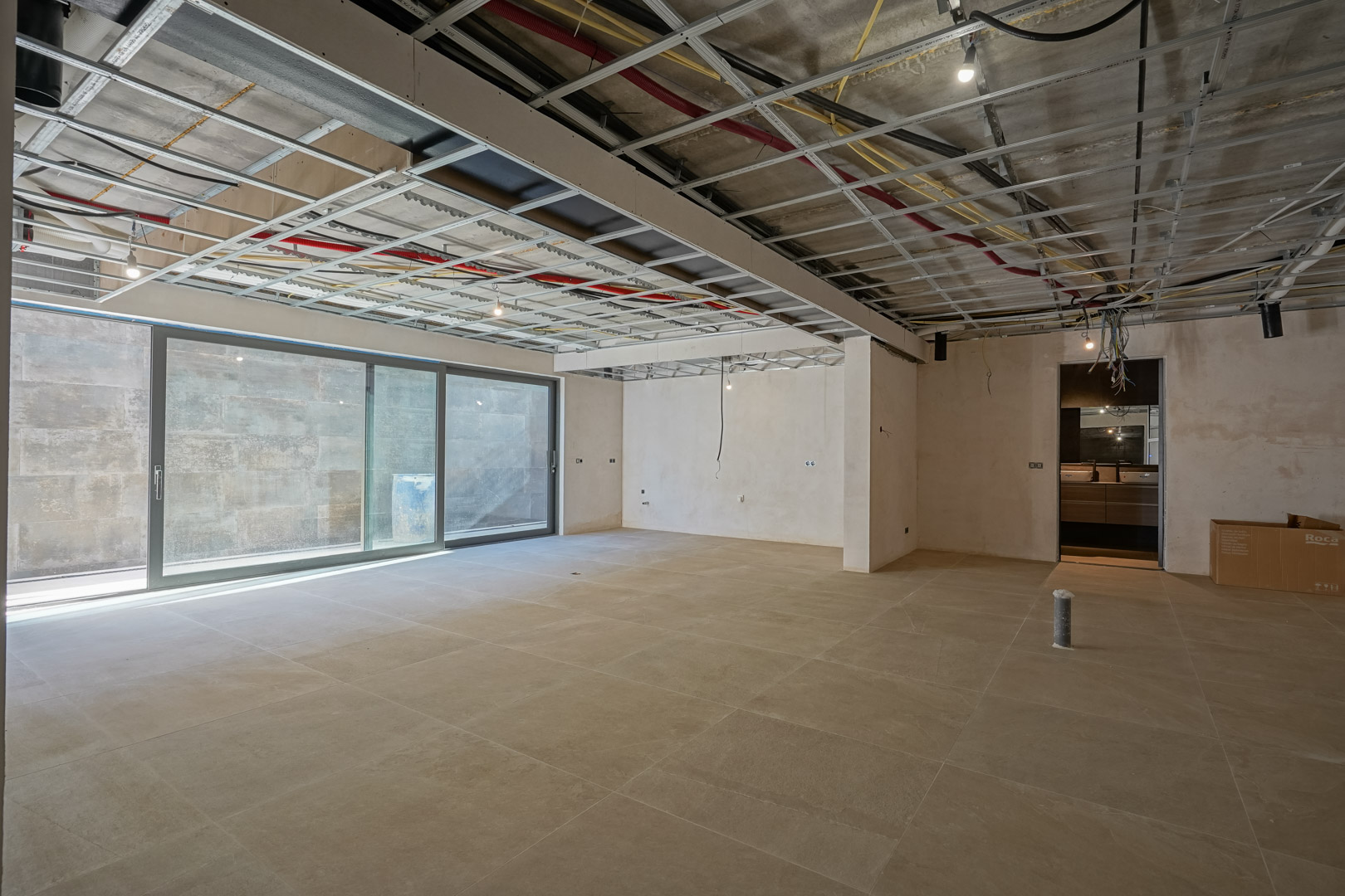 Sous-sol spacieux en cours de finition avec carrelage grand format, baie vitrée coulissante et structure de plafond technique apparente.