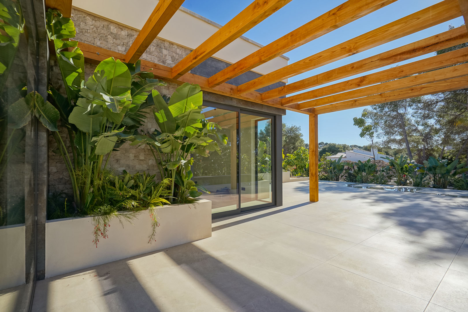 Terrasse extérieure avec pergola en bois d'iroko, carrelage grand format et jardinière intégrée avec plantes tropicales devant les baies vitrées.