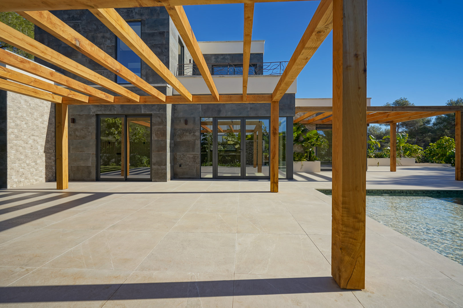 Terrasse extérieure avec pergola en bois d'iroko et carrelage clair, située à côté de la piscine et d'une façade moderne en pierre grise.