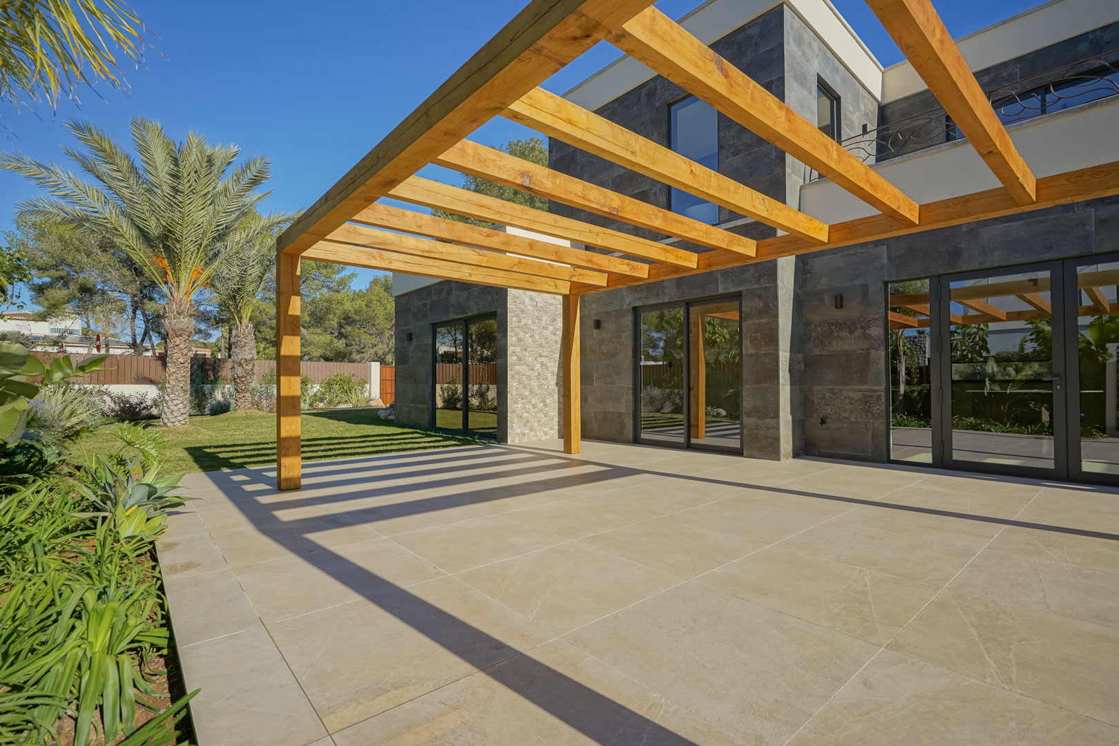 Terrasse extérieure avec pergola en bois d'iroko et carrelage grand format, bordée par une façade en pierre et un jardin méditerranéen soigné.
