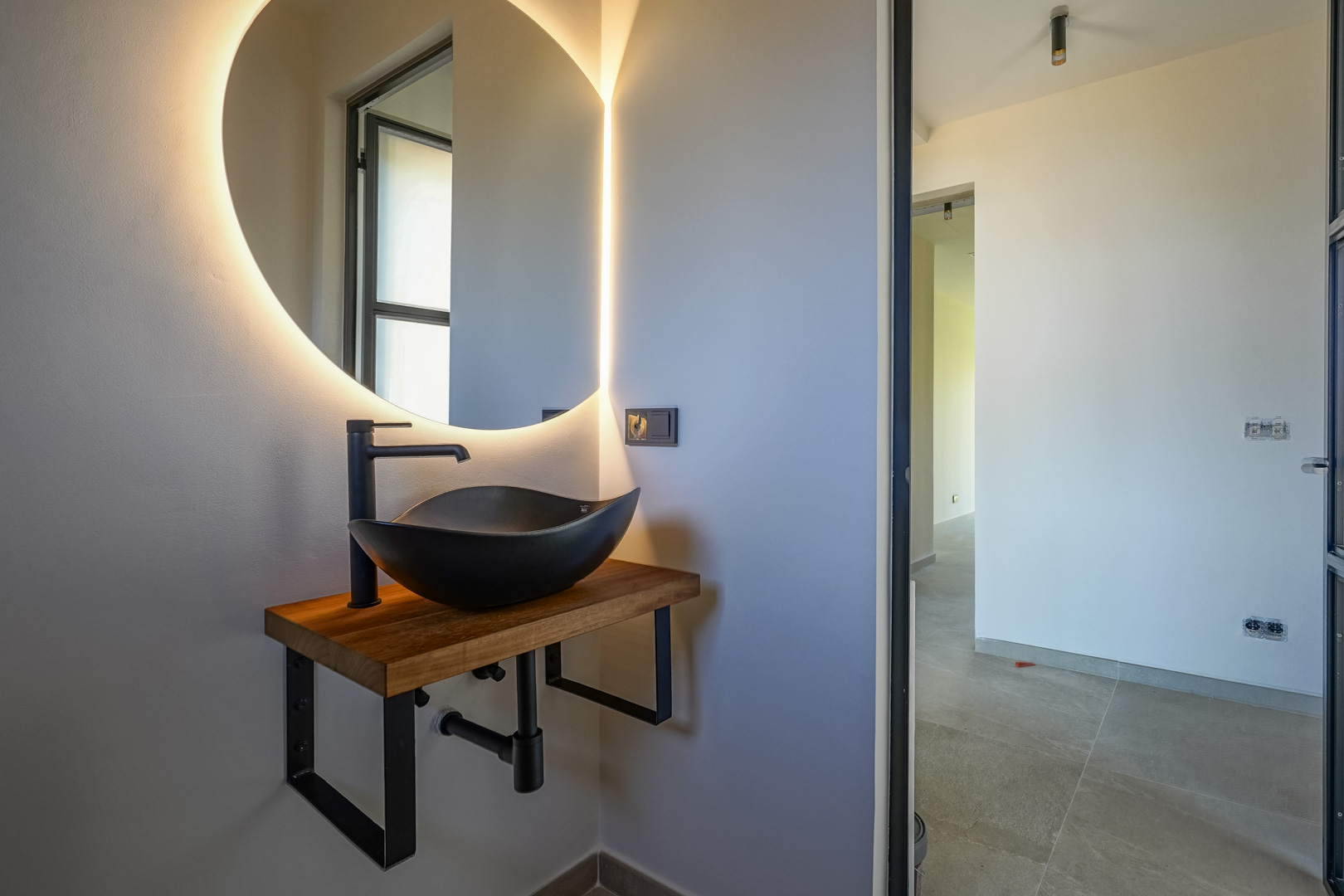 Toilettes invités avec vasque à poser noire sur plan en bois massif, miroir circulaire rétroéclairé par LED et robinetterie noire mate.