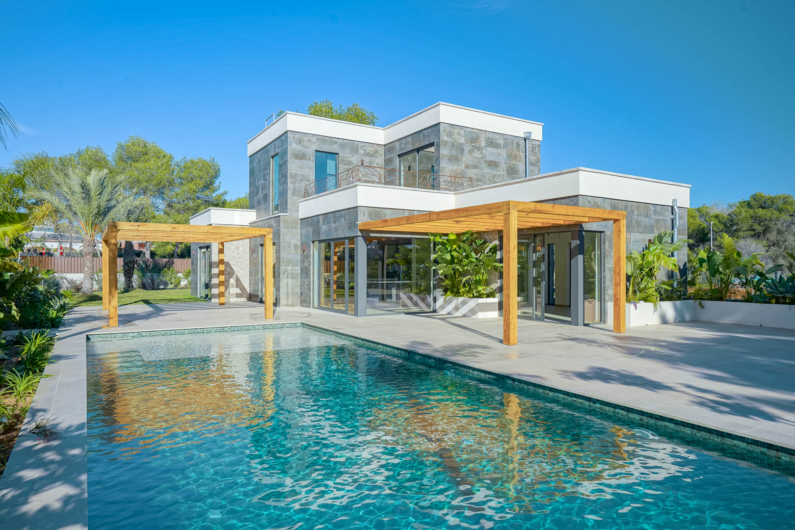 Vue extérieure de la villa avec piscine de style Bali, terrasses spacieuses et pergolas en bois d'iroko devant une façade moderne en pierre naturelle.