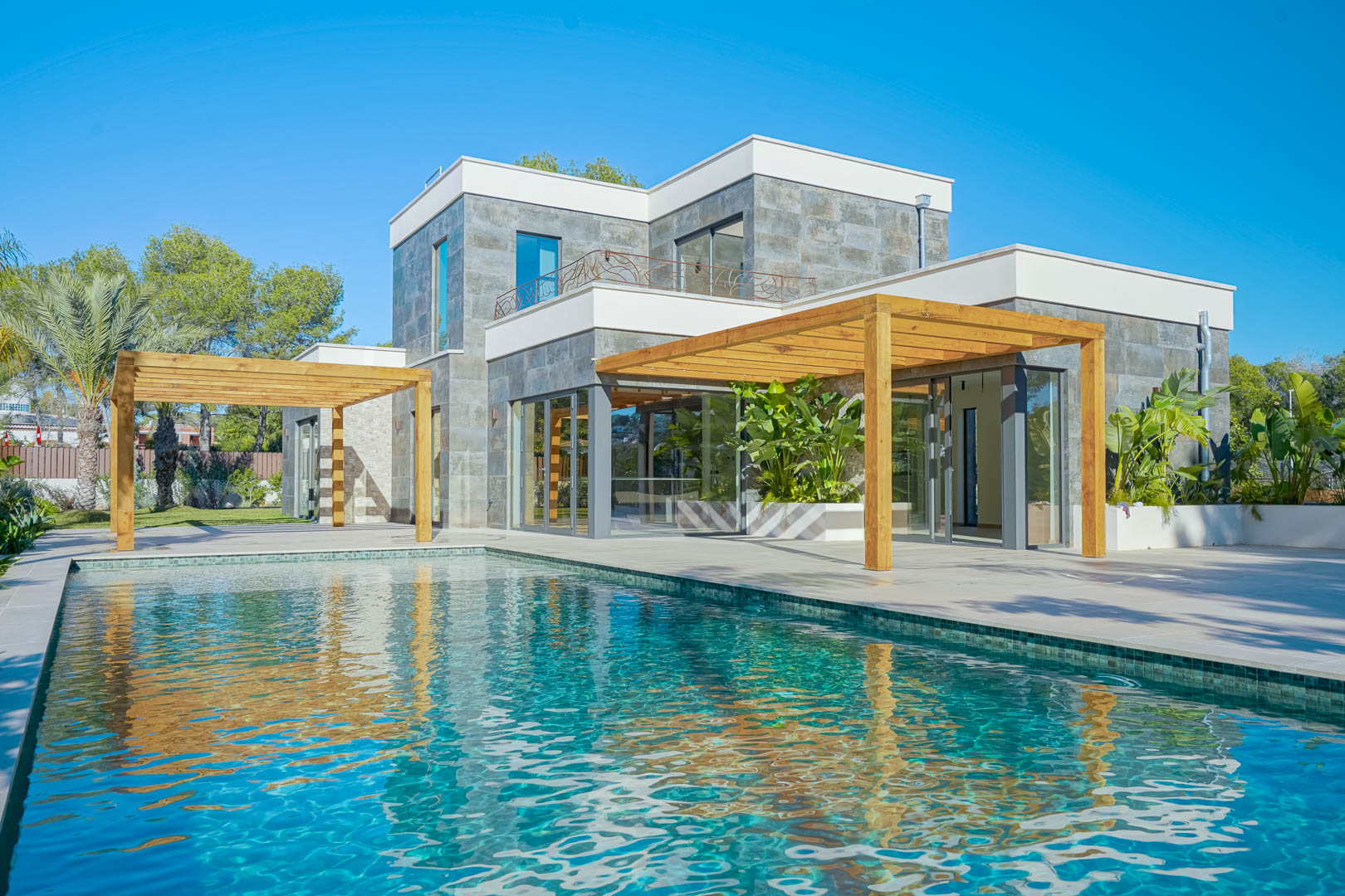 Vue extérieure de la villa avec piscine en mosaïque, pergolas en bois d'iroko et façade en pierre naturelle avec de grandes baies vitrées.