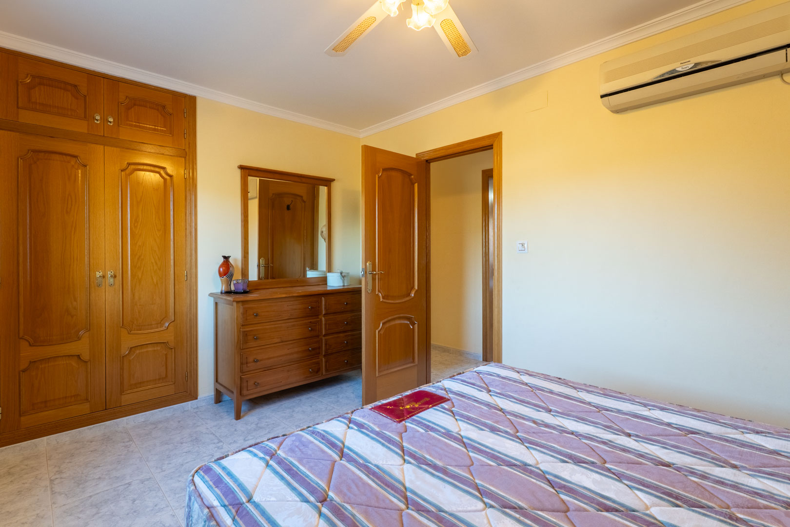 Chambre avec placards intégrés en bois, commode assortie avec miroir, climatisation réversible et ventilateur de plafond sur sol carrelé clair.