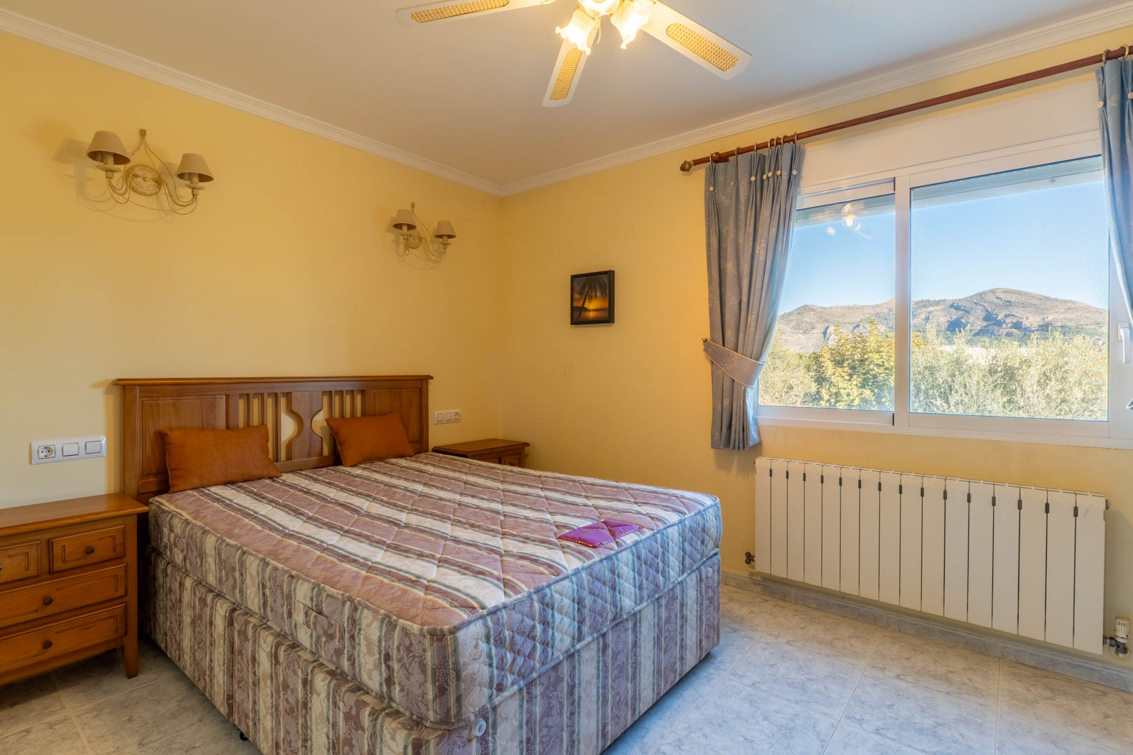 Chambre lumineuse avec mobilier en bois classique, ventilateur de plafond et grande fenêtre offrant une vue dégagée sur les montagnes environnantes.